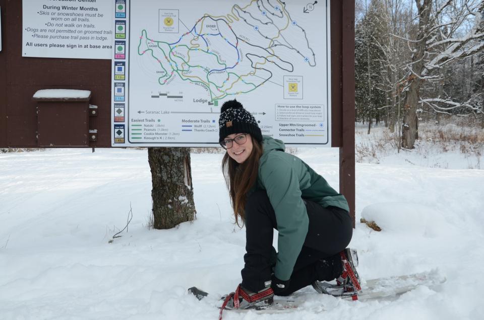 Girl smiles at the camera as she tightens her snowshoes in front of a Dewey Mountain sign