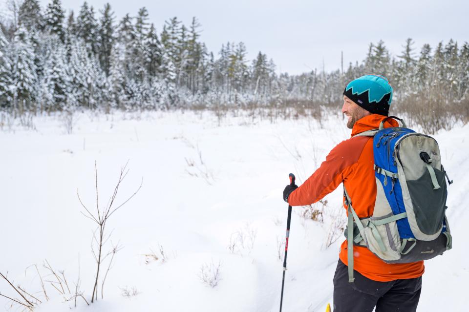 A man in an orange hoodie looks at the view while crosscountry skiing.