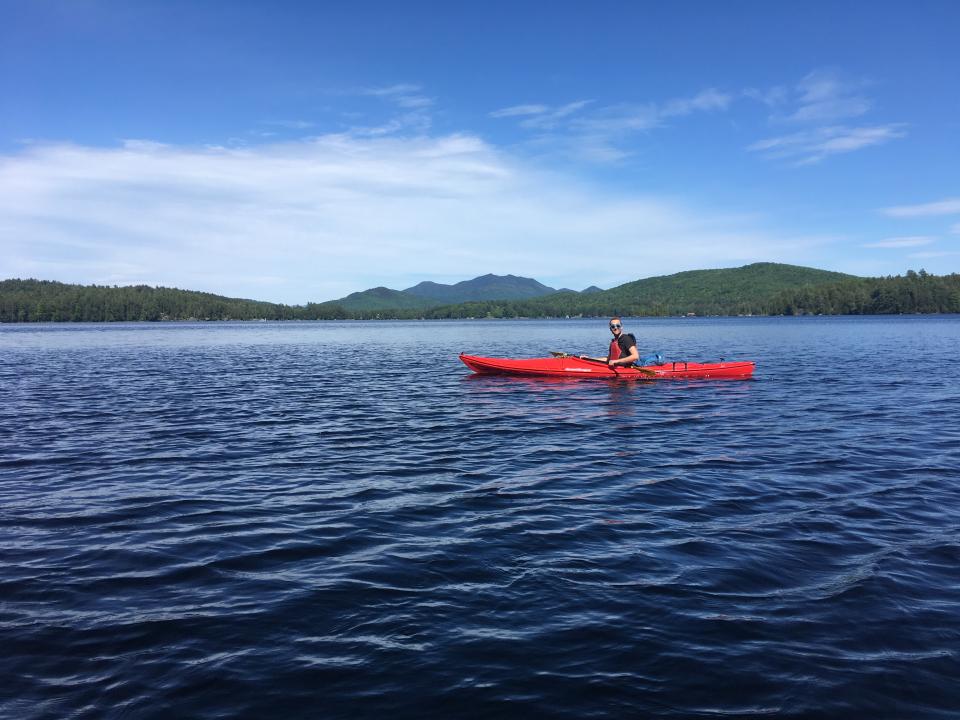 Kayaker in a red kayak paddling across Lower Saranac Lake, with mountains and blue skies in the background
