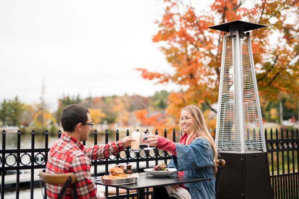 A man and woman toast drinks at an outdoor restaurant table, with vibrant fall foliage in the background.