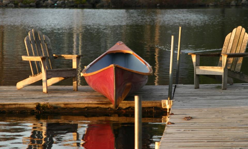 A canoe sits on top of a dock between two Adirondack chairs on a fall day.