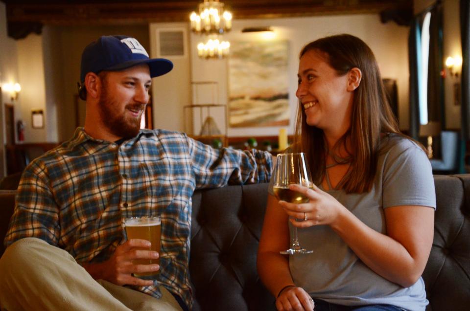 A couple smiles at each other while enjoying a beer and wine as they sit on a couch
