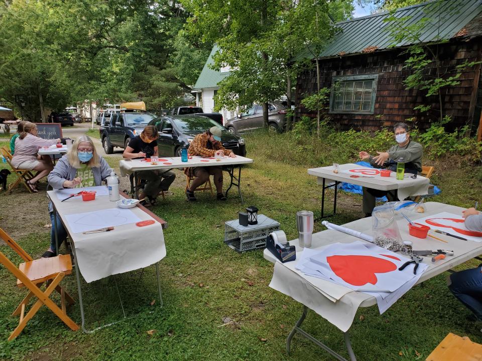 People sit at tables in a yard outdoors and paint hearts on fabric for the Heart Banner Project.