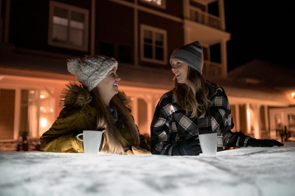 Two women laugh over hot cocoa in front of a hotel at night.