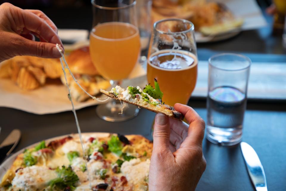 two hands pull cheese apart from a flatbread pizza with beer in the background.