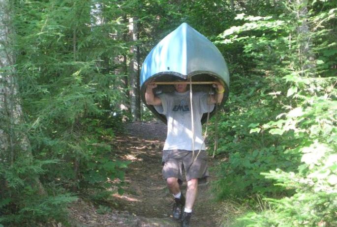 A man walks through the woods with a canoe held above his head.