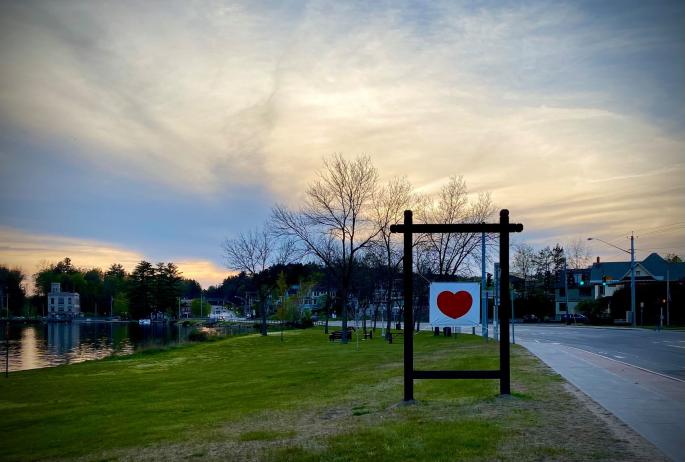 A poster with a red heart is displayed on a lawn with the sun setting in the background
