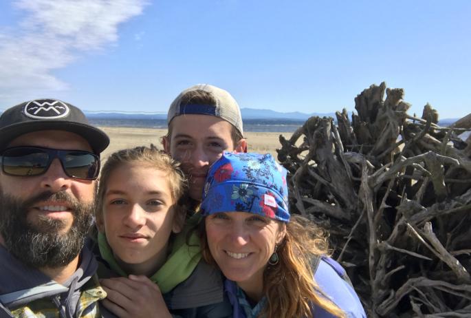 Jason Smith stands with his wife and two children on a beach with a large pile of driftwood behind them