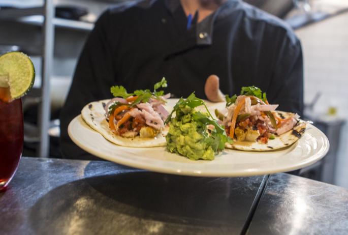 Two tacos sit on a plate prepared in a chef's hand.