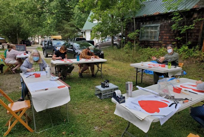 People sit at tables in a yard outdoors and paint hearts on fabric for the Heart Banner Project.