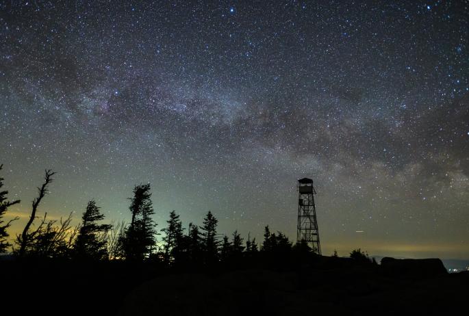 A nighttime image of the fire tower on Hurricane Mountain, with the Milky Way glowing bright against the dark sky