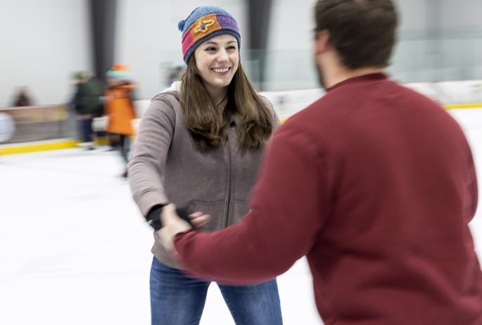 A man and woman laugh as they skate holding hands.