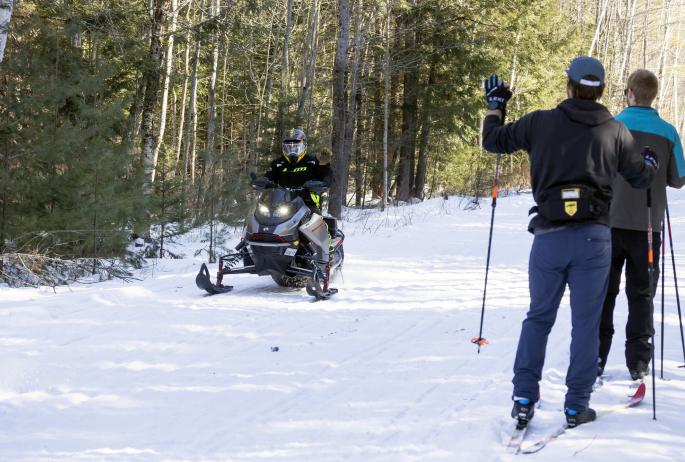 a snowmobiler passes two cross-country skiers on a snow-covered trail.