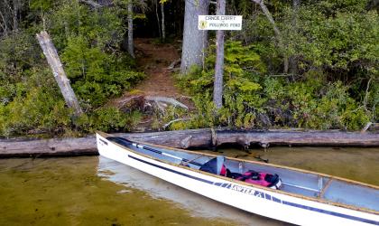 Canoe floats in the water with gear near the shore. Polliwog sign in the background attached to a tree.