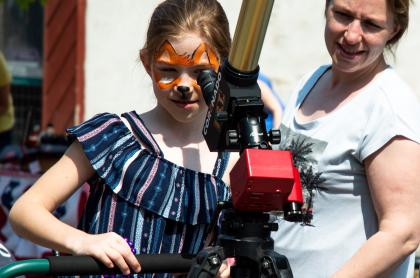 Kid with butterfly face painting looks through a telescope during the daytime.