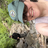 A woman wearing a baseball cap smiles in front of a rocky trail.