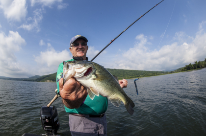 A man holds a large mouth bass with a fishing pole with the water and mountain in the background