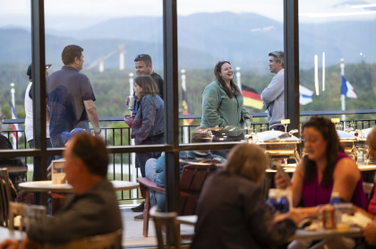 People seen mingling inside and outside of big glass windows with view of Lake Placid from outside deck