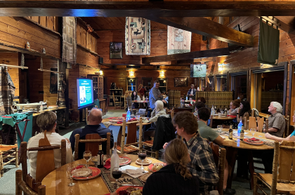 A group of people seated at round tables in a rustic wooden lodge listen to a presentation shown on a screen.