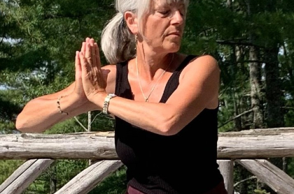 A woman holds a yoga pose with her eyes closed outside surrounded by pine trees.
