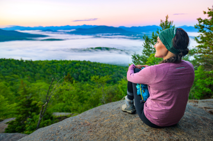 Woman in a purple shirt sits on the edge of Baker Mountain and stares out overtop of the green trees below with light cloud cover and other Adirondack mountains in the distance