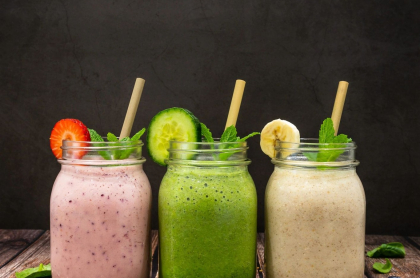 Three colorful mason jars of fresh cold pressed juice on a table surrounded by mint leaves and blueberries.