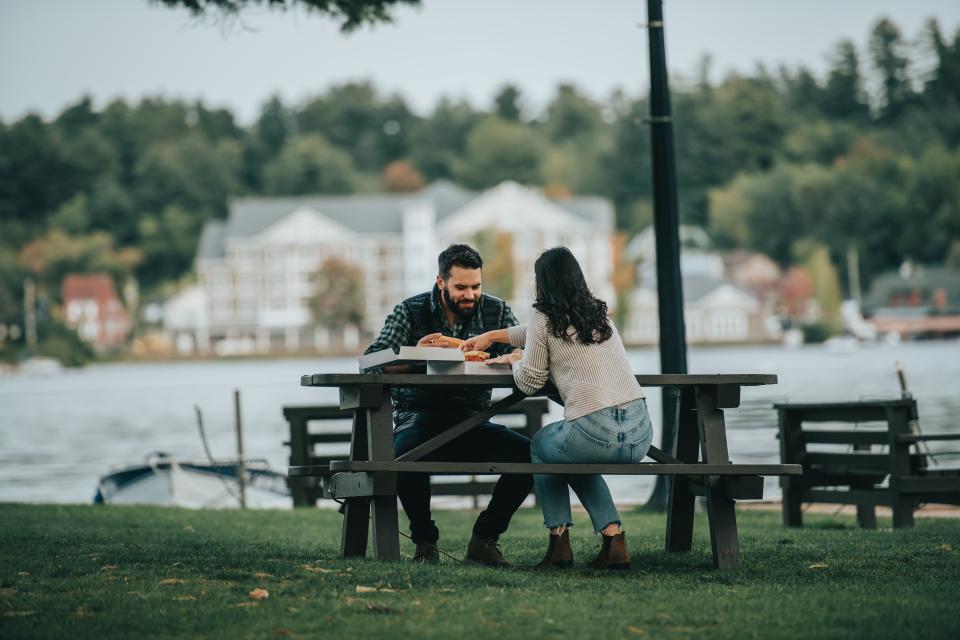 A couple enjoys a meal at a picnic table with the Saranac Waterfront Lodge in the background
