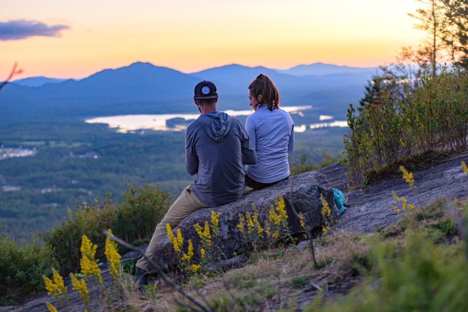 A man and woman sit on a mountain summit in the warm colors of a spring sunset.
