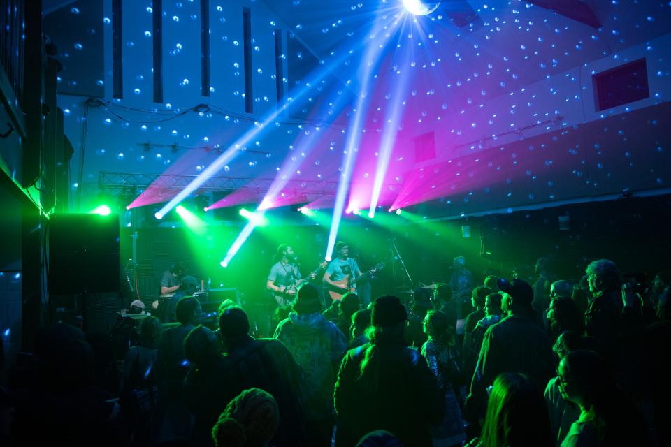 A band plays in an open room with colorful lights of pink and green.