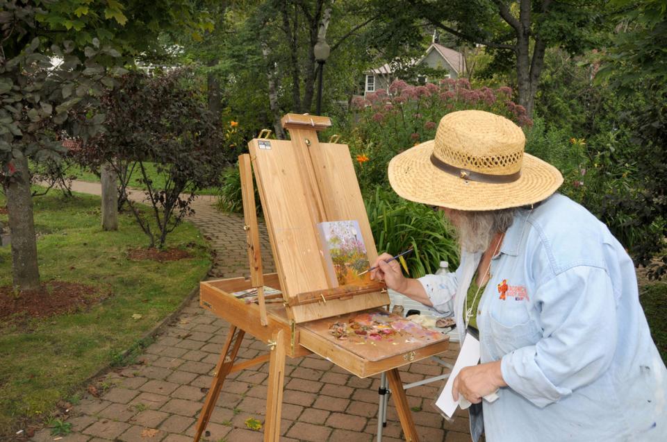 A person in a straw hat paints at an easel in a park, surrounded by greenery.