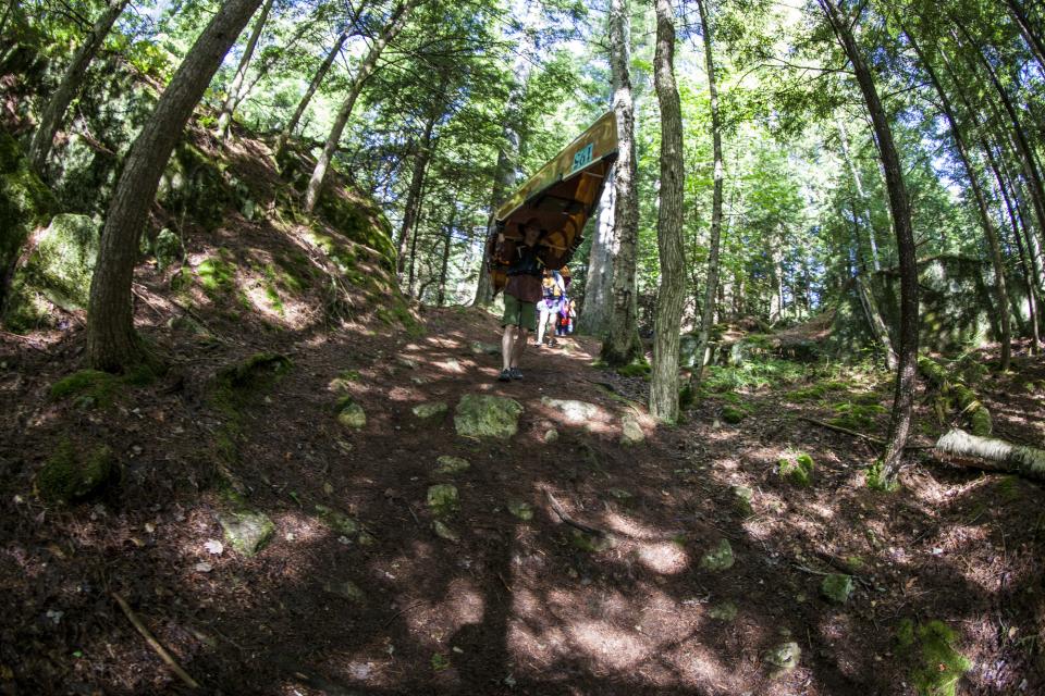 A man carries a yellow canoe over his head on a dirt trail through the woods