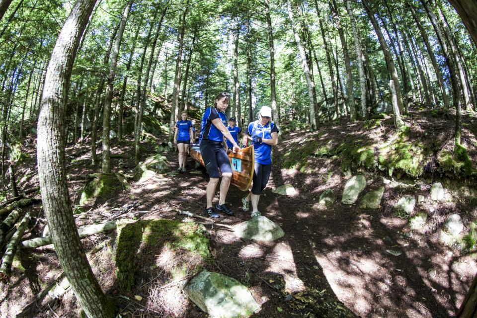 A team of women in matching blue shirts carry a beautiful wooden canoe on a dirt path through the woods to the next launching point in the Adirondack 90-Miler
