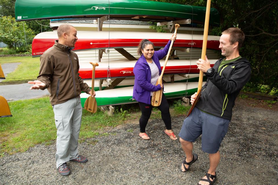 A couple uses canoe oars as air guitars in front of a rack of canoe rentals while a Saranac Lake outfitter explains the rental options available.