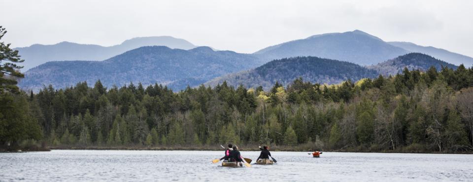 Two canoes and a kayak paddle away from the camera and towards the backdrop of the Adirondack mountains