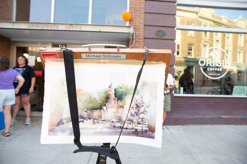 An artist's easel holds a painting of Main Street in Saranac Lake with shops behind it