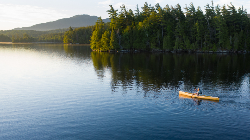A man paddles a solo canoe during sunrise with mountains in the background.