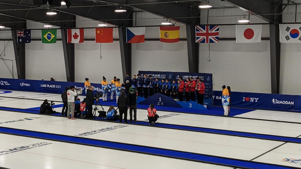 Athletes stand by a podium next to an iced curling rink.