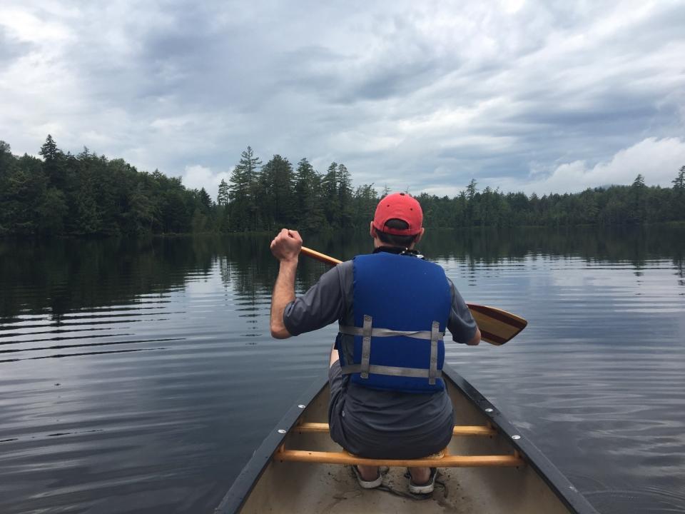 A man with a blue life vest and red hat paddles from the front of a canoe on a pond surrounded by evergreens.