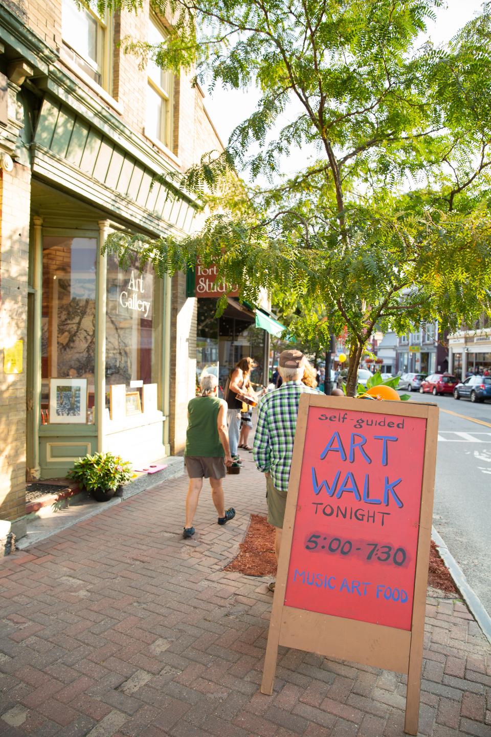 A colorful Art Walk sign stands before a busy sidewalk with artists and visitors.