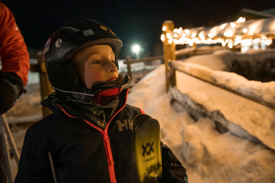 A young boy stands with his downhill skis and helmet next to an illuminated fence.