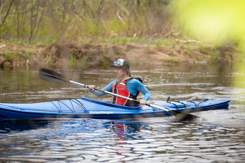 A woman in a blue kayak paddles down a river amid bright green spring vegetation.