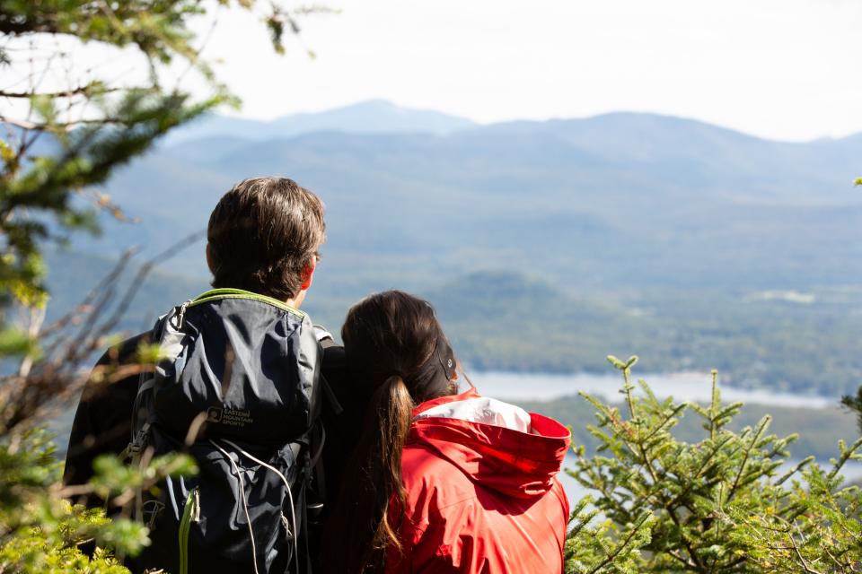 Two hikers enjoy a view from a mountain with distant mountains and a lake in the background.
