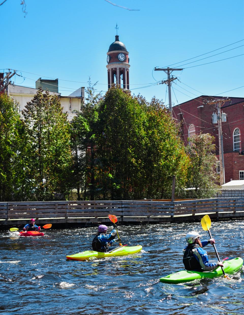 A group paddles their kayaks on a river with the town in the background.