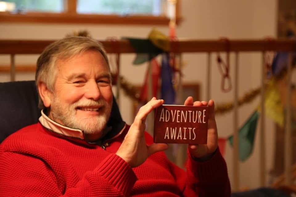 An elderly man, Tom Boothe, in a red sweater smiles and holds a small sign reading "Adventure Awaits,"