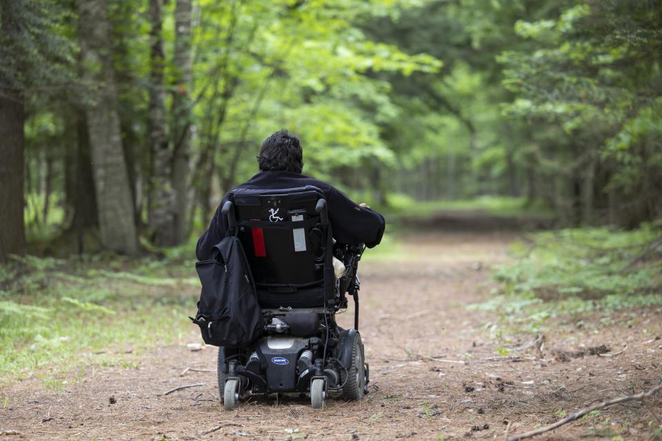 A man in a power wheelchair moves down an attractive, flat earthen path in a green forest.