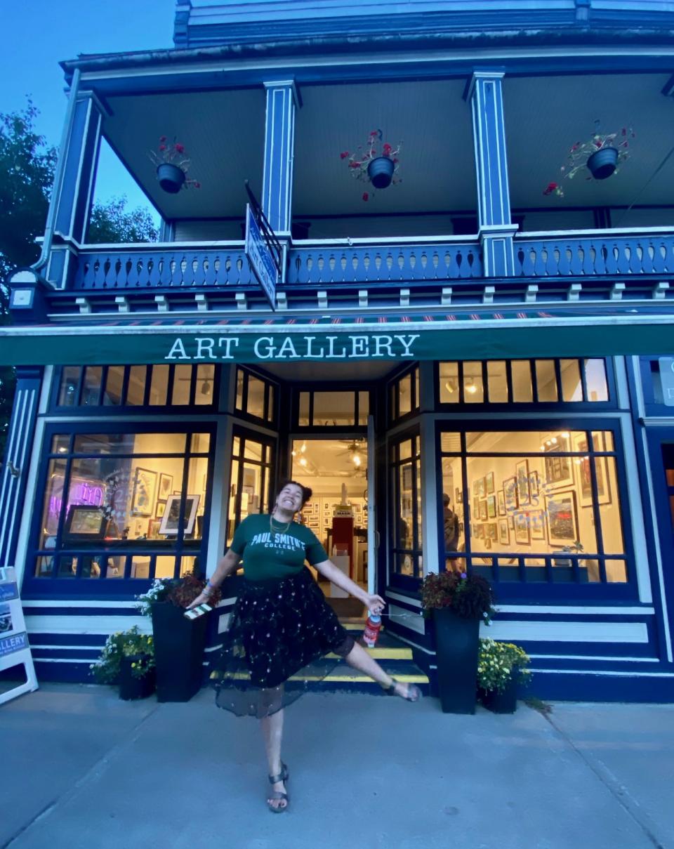 The exterior of a charming, brightly lit art gallery with awning over the door. A college student poses happily outside the door.
