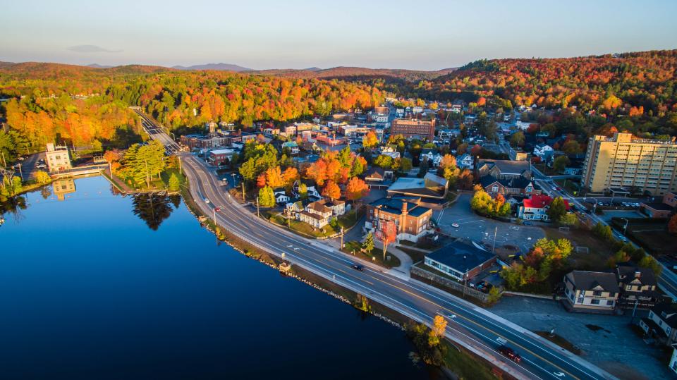 An aerial shot of downtown Saranac Lake. Fall foliage, lakes, buildings, and the highway are featured.