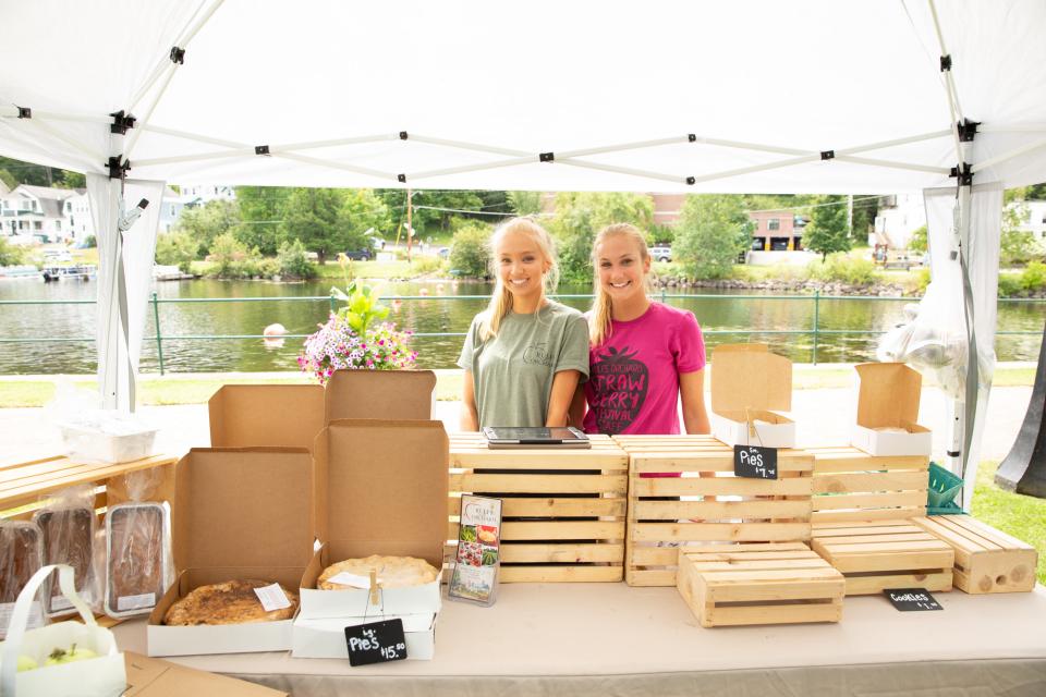 Two girls stand behind their products at the farmers market. Lake Flower and houses serve as the background.