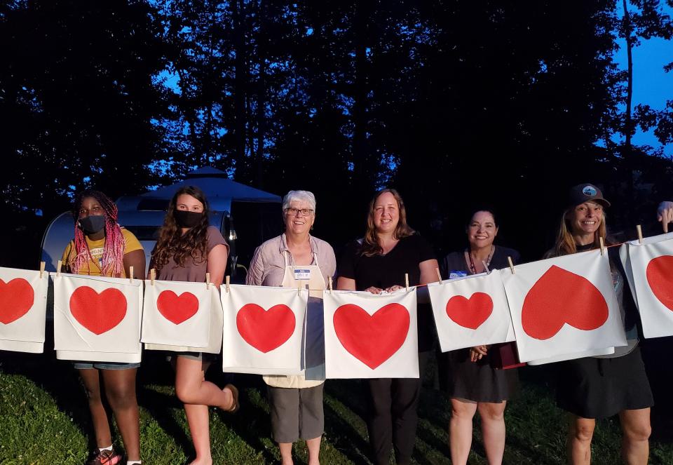 Six women stand behind a clothesline that holds a number of red hearts printed on white fabric.