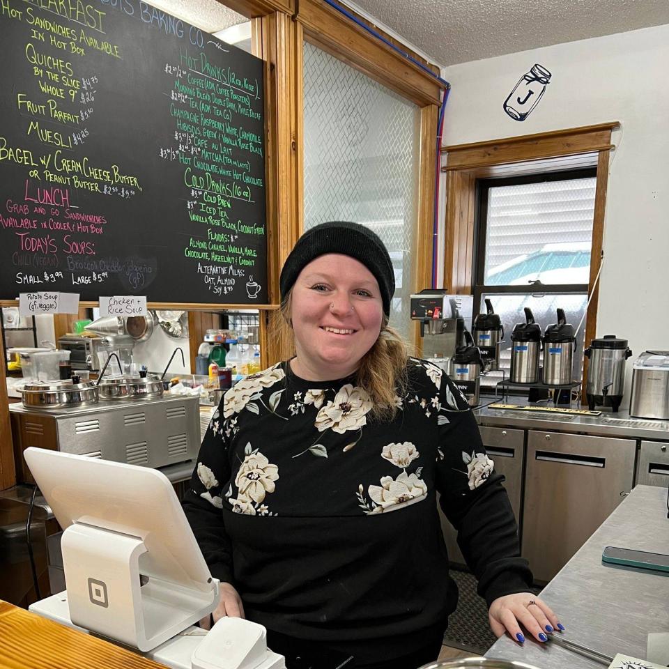 A woman smiling behind a counter, serving at a local cafe with a menu in the background